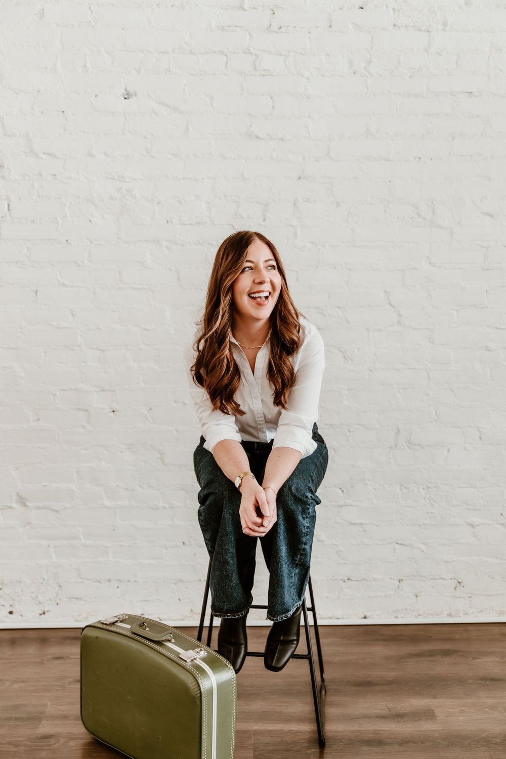 Candid lifestyle portrait of someone sitting on a chair laughing next to vintage olive green luggage against white wall.
