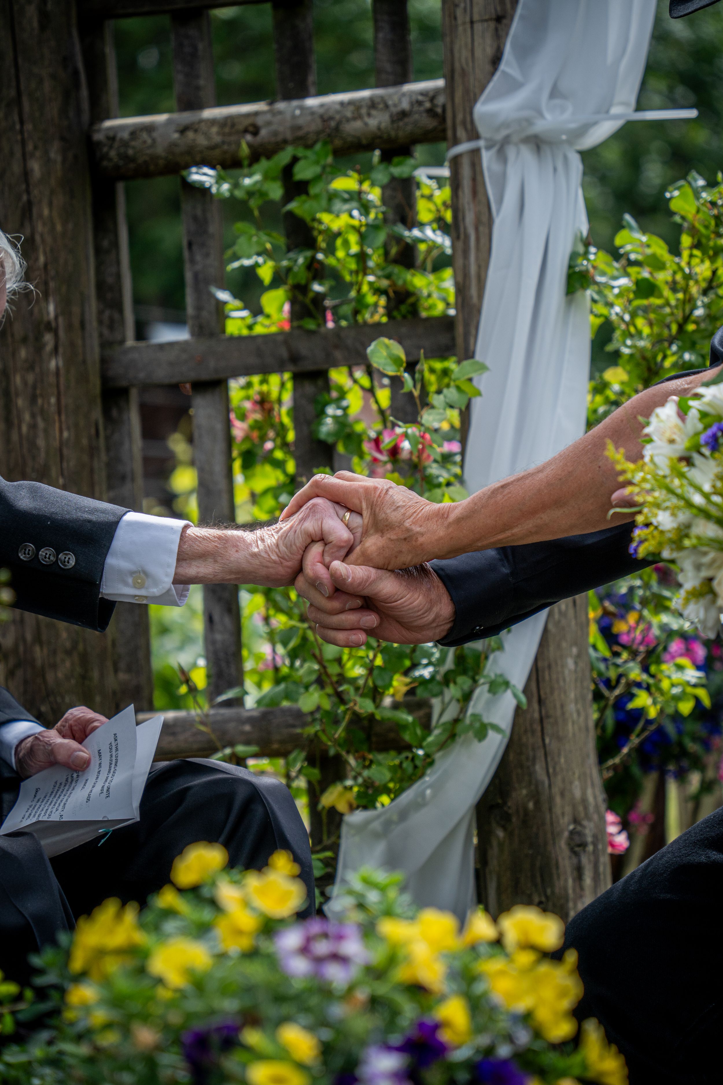 Two hands clasped in ceremony amongst colorful garden flowers and white fabric decor.