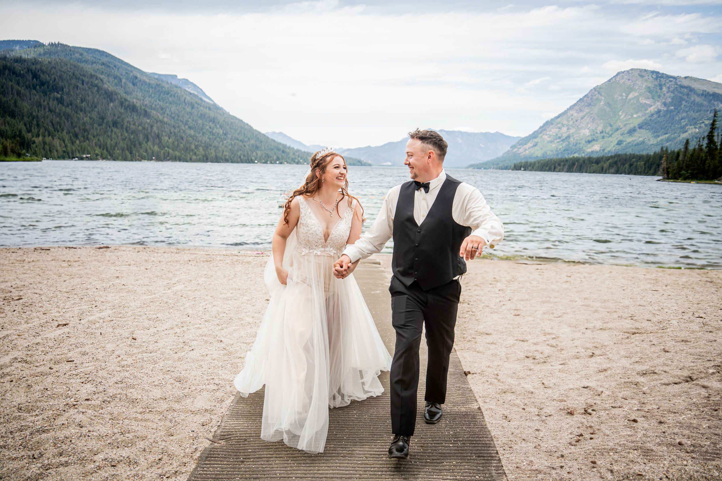 A couple in wedding attire walks along a scenic mountain lake shore with dramatic peaks in the background.