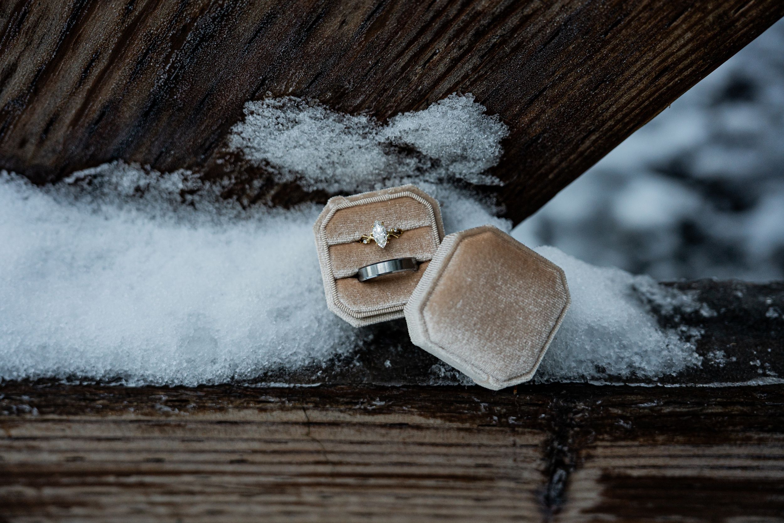 Two diamond engagement rings displayed in a velvet hexagonal ring box resting on snow-dusted rustic wood.