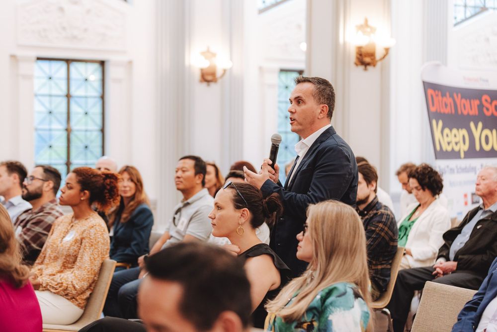 An audience member stands to ask a question during a conference session in an elegant room with wall sconces.