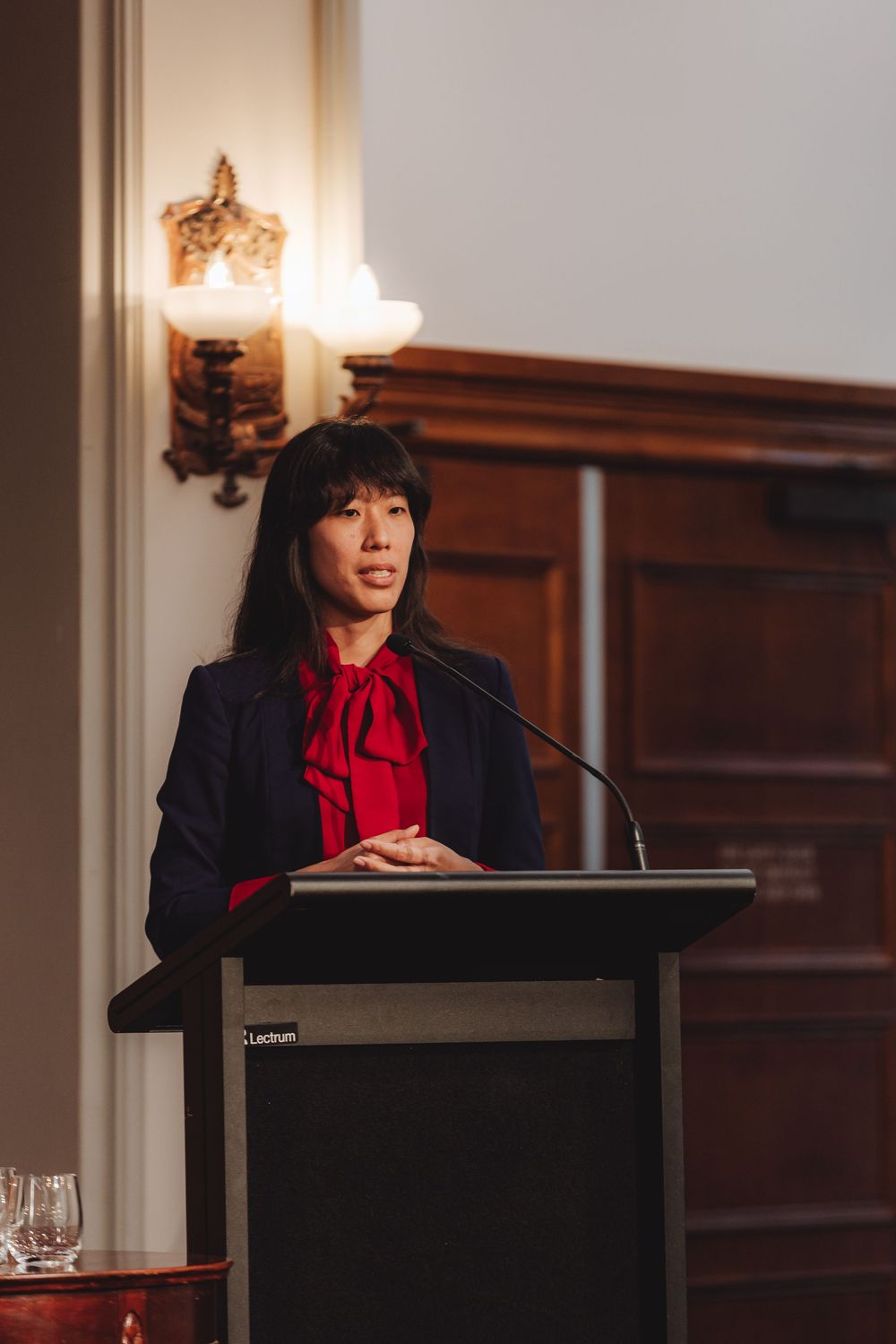 A speaker stands at a podium in a formal setting with wood paneling and warm lighting.