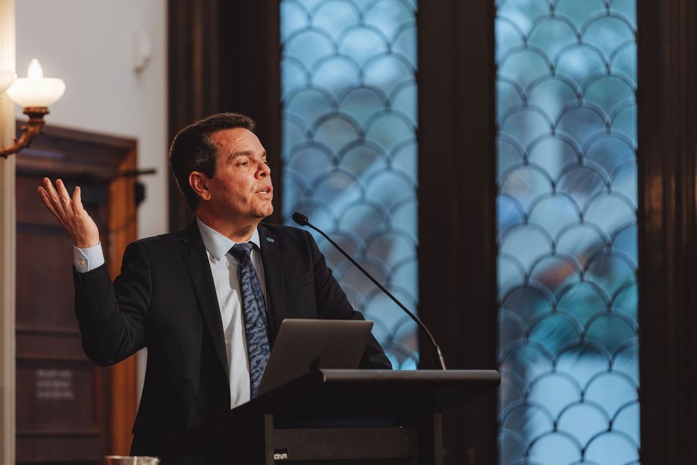 A speaker gestures while presenting at a wooden podium in an elegant venue with decorative windows.