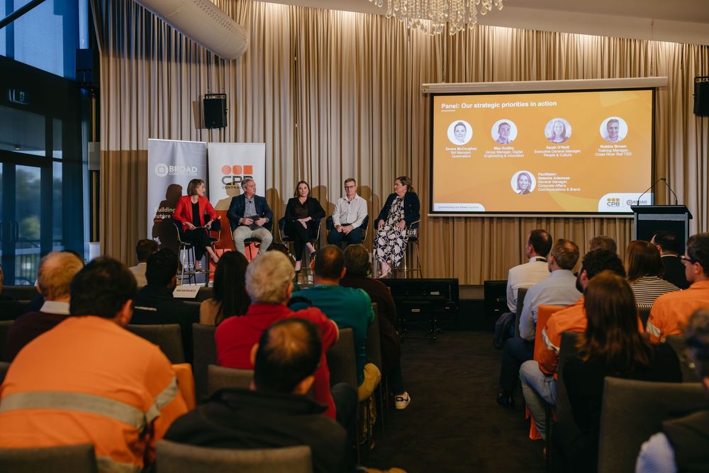 Panel discussion event with speakers on stage and audience members wearing high-visibility orange safety vests.