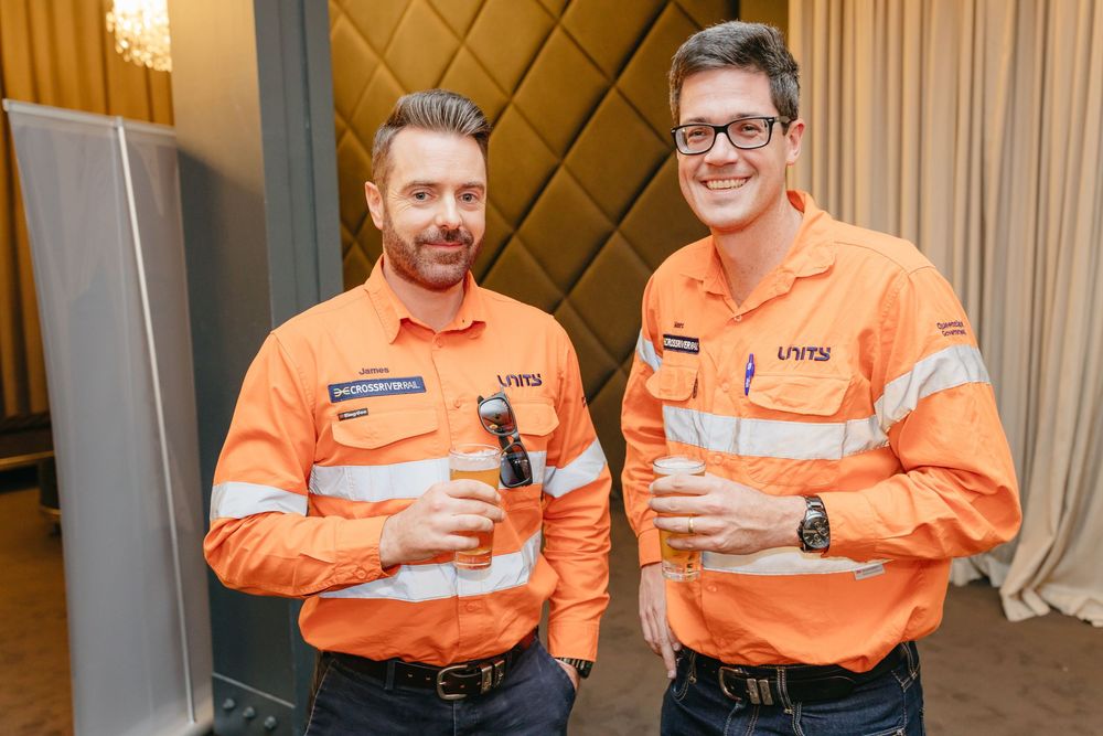 Two workers in bright orange safety uniforms holding beverages at an indoor event.