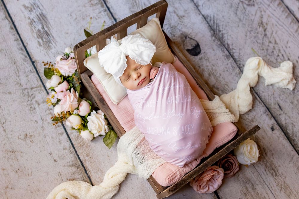 A small wooden crate decorated with flowers holds a sleeping baby in pink swaddle on a rustic wooden floor.