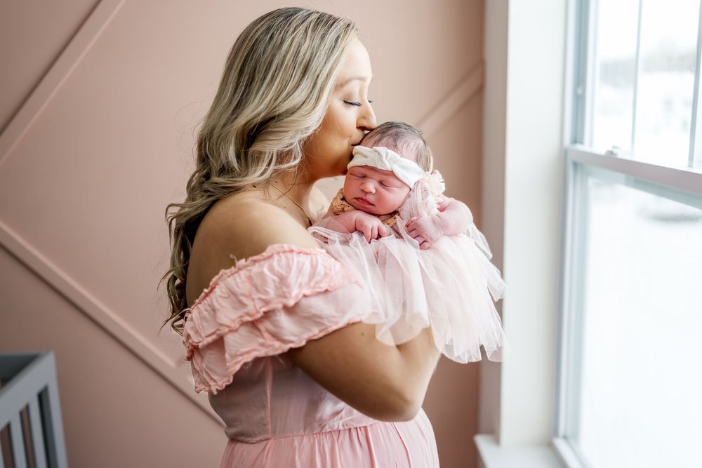 A mother and baby in matching pink dresses share tender moments by a window in a soft, naturally lit room.