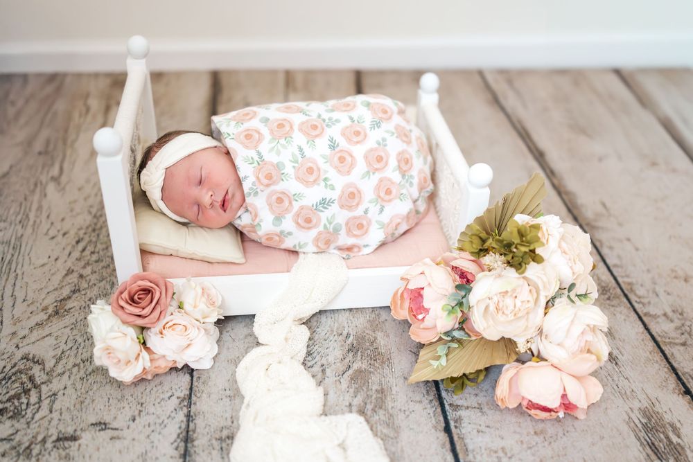 Newborn sleeping on white wooden prop bed with pink roses on rustic wood floor.