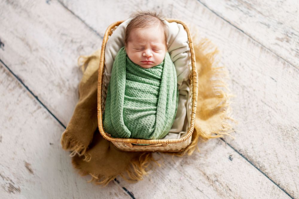 A sleeping newborn wrapped in soft green swaddle cloth lies on a wooden floor with cream-colored fabric accents.