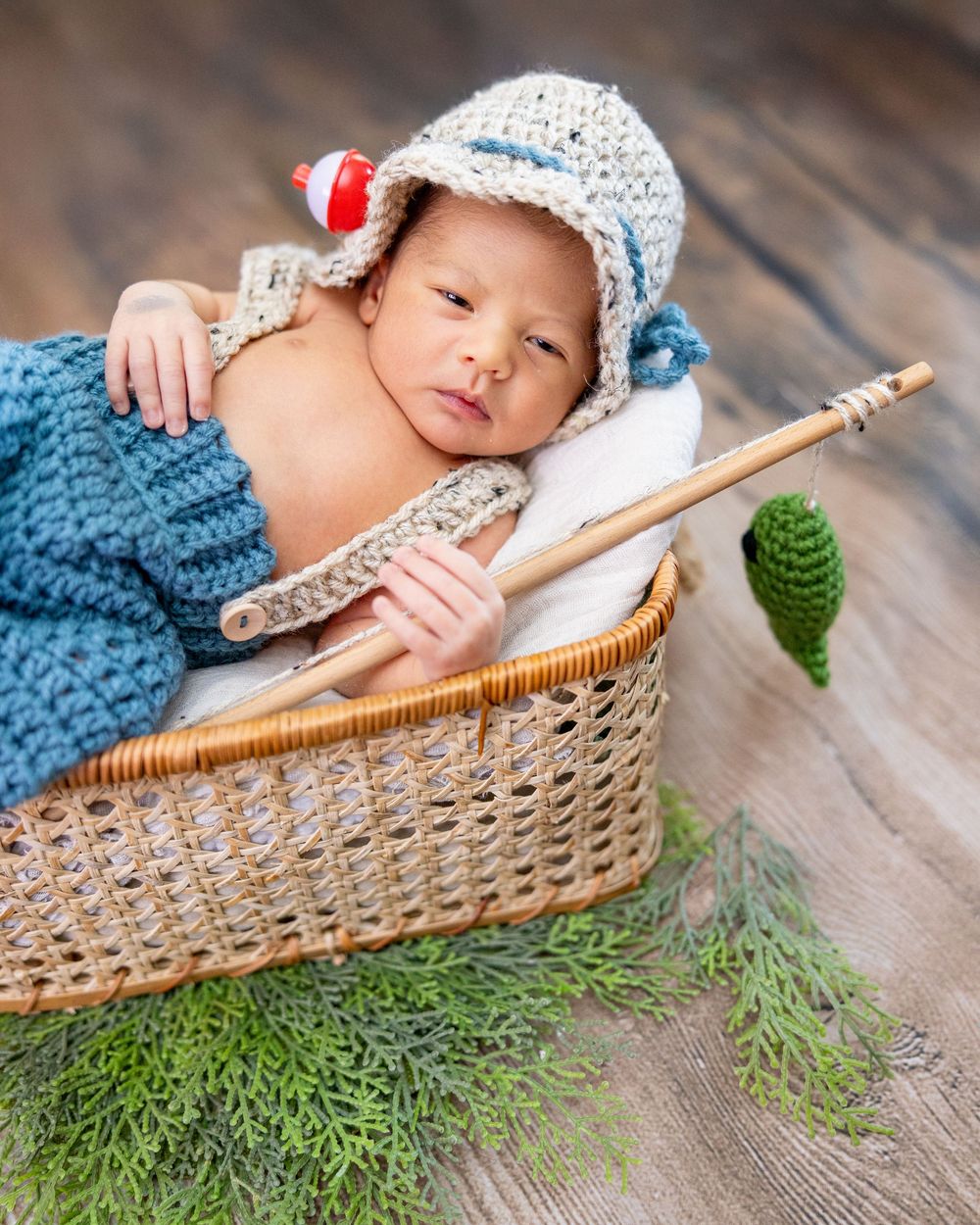 A sleeping baby wearing a knit hat rests peacefully in a woven basket with evergreen sprigs on wooden flooring.