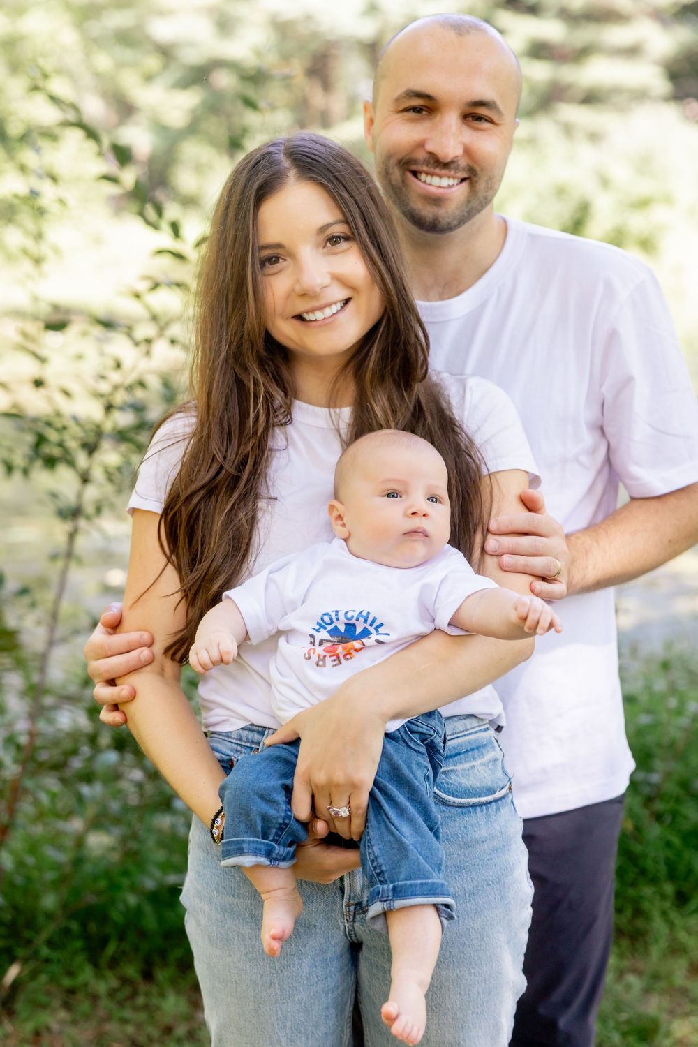 A family in matching white t-shirts and jeans poses outdoors with their baby in a sunny natural setting.