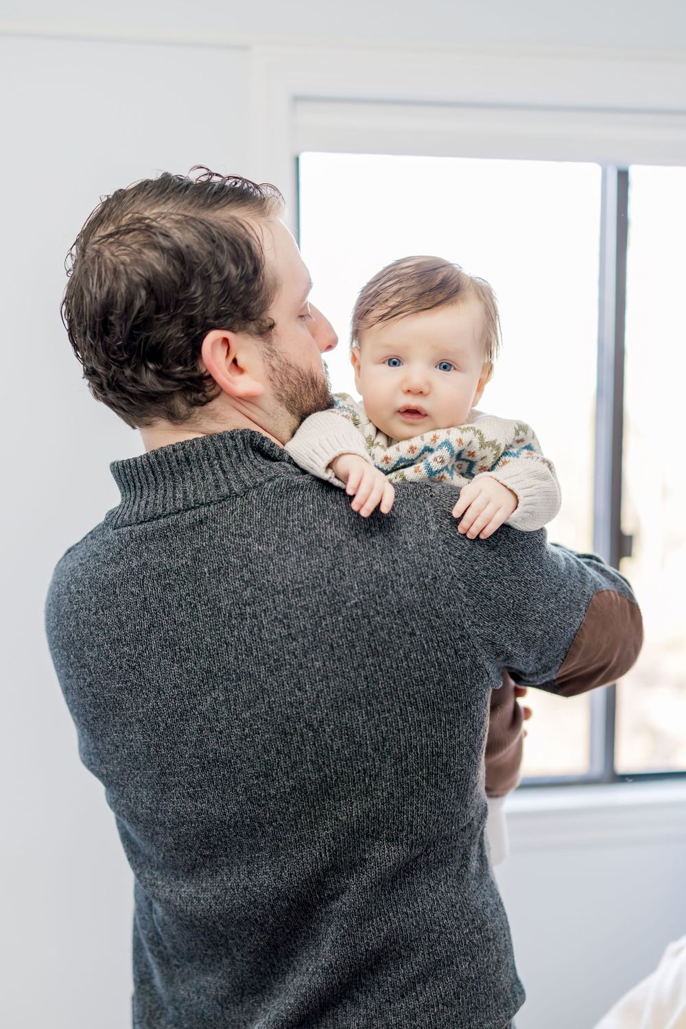 A person wearing a gray sweater holds a baby while looking out a bright window in a sequence of three photos.