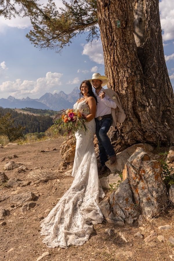 A couple poses for a wedding photo against a majestic mountain backdrop near a large tree trunk on a dirt path.