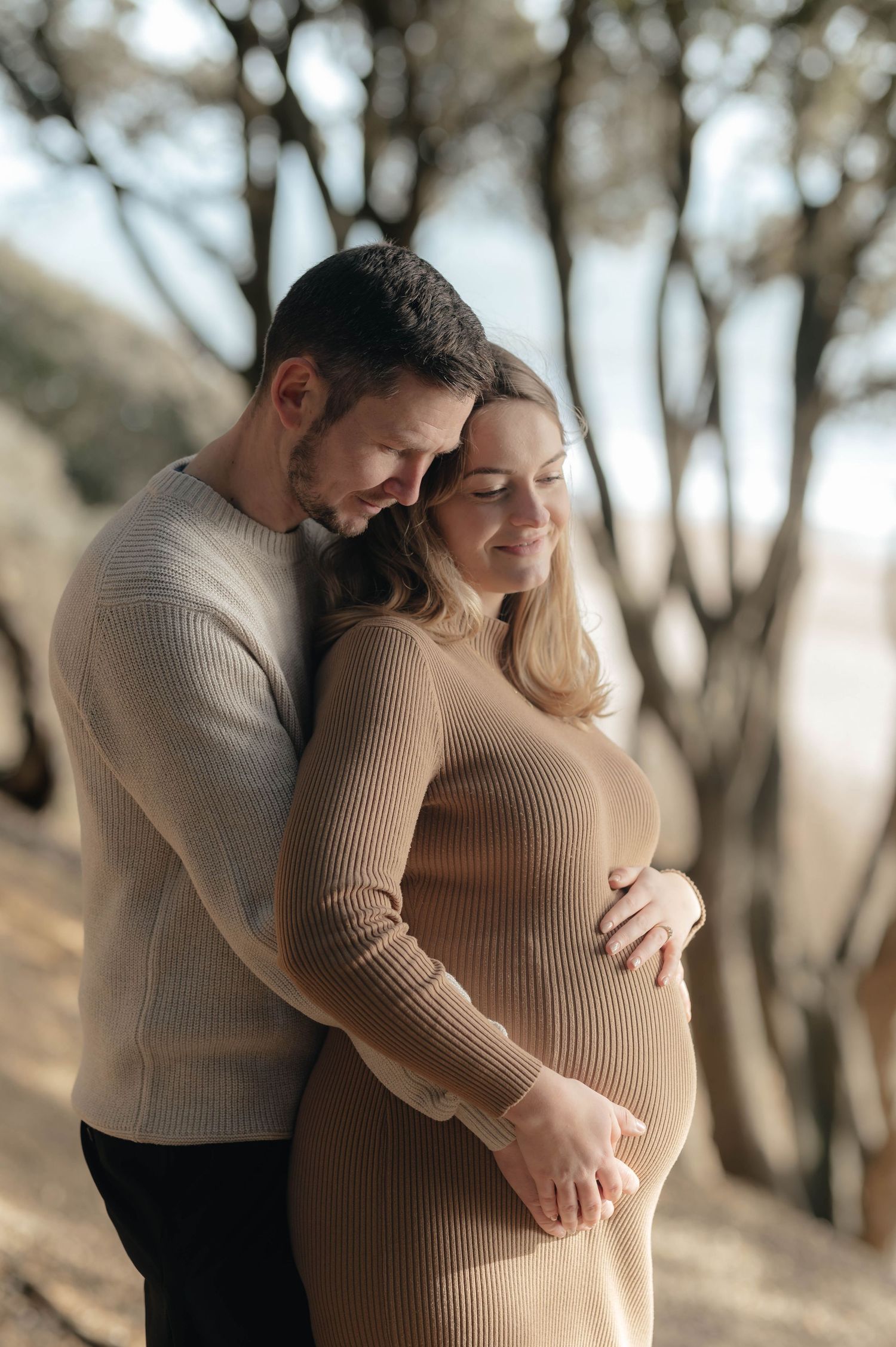 A couple shares an intimate moment outdoors while holding a pregnant belly wearing a blue dress and black quilted jacket.