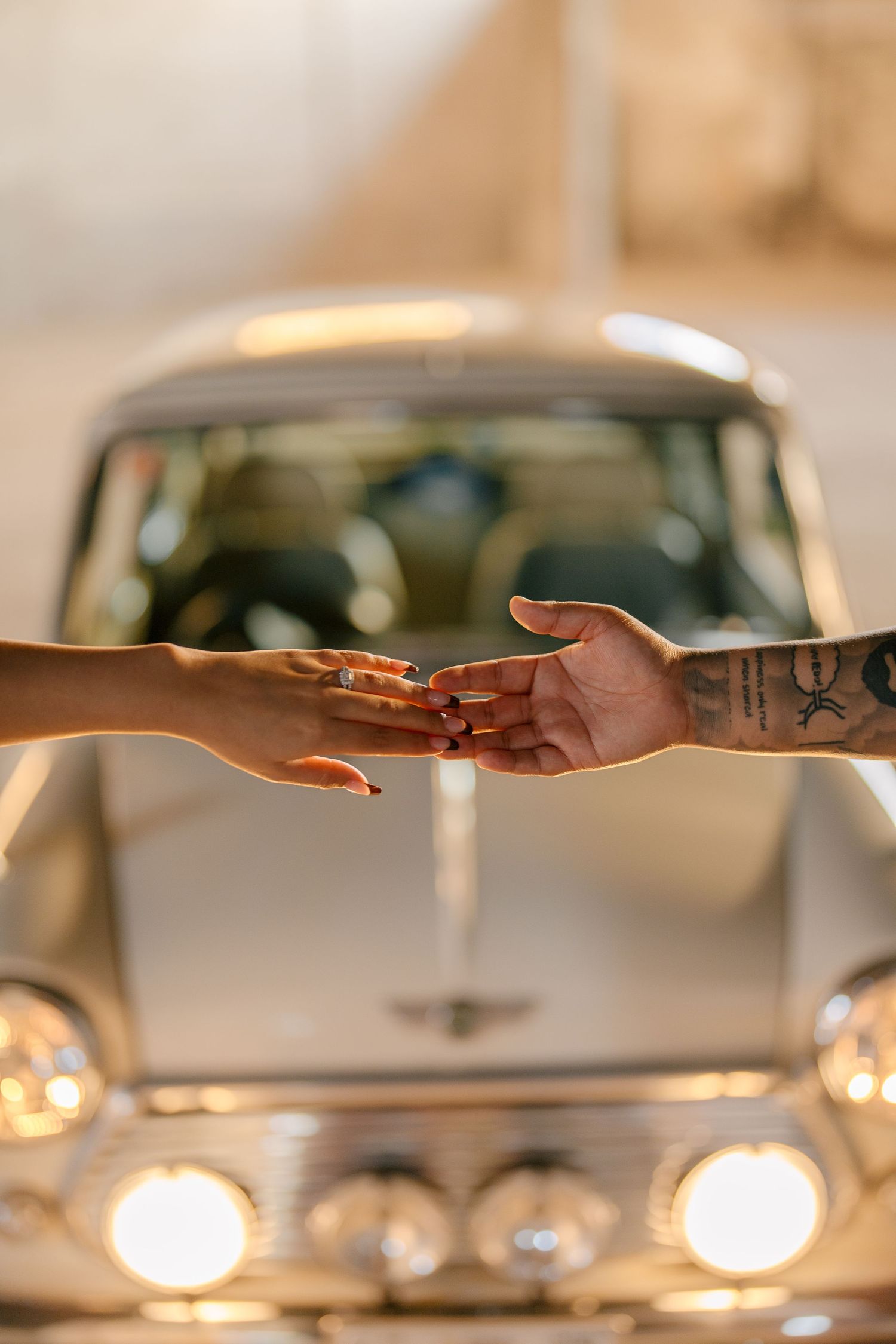 Close-up of hands reaching toward each other in front of vintage car with glowing headlights.
