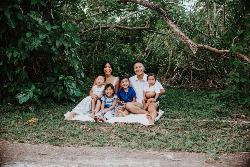 Family of six poses on a blanket outdoors, surrounded by lush green trees, smiling together for a portrait.