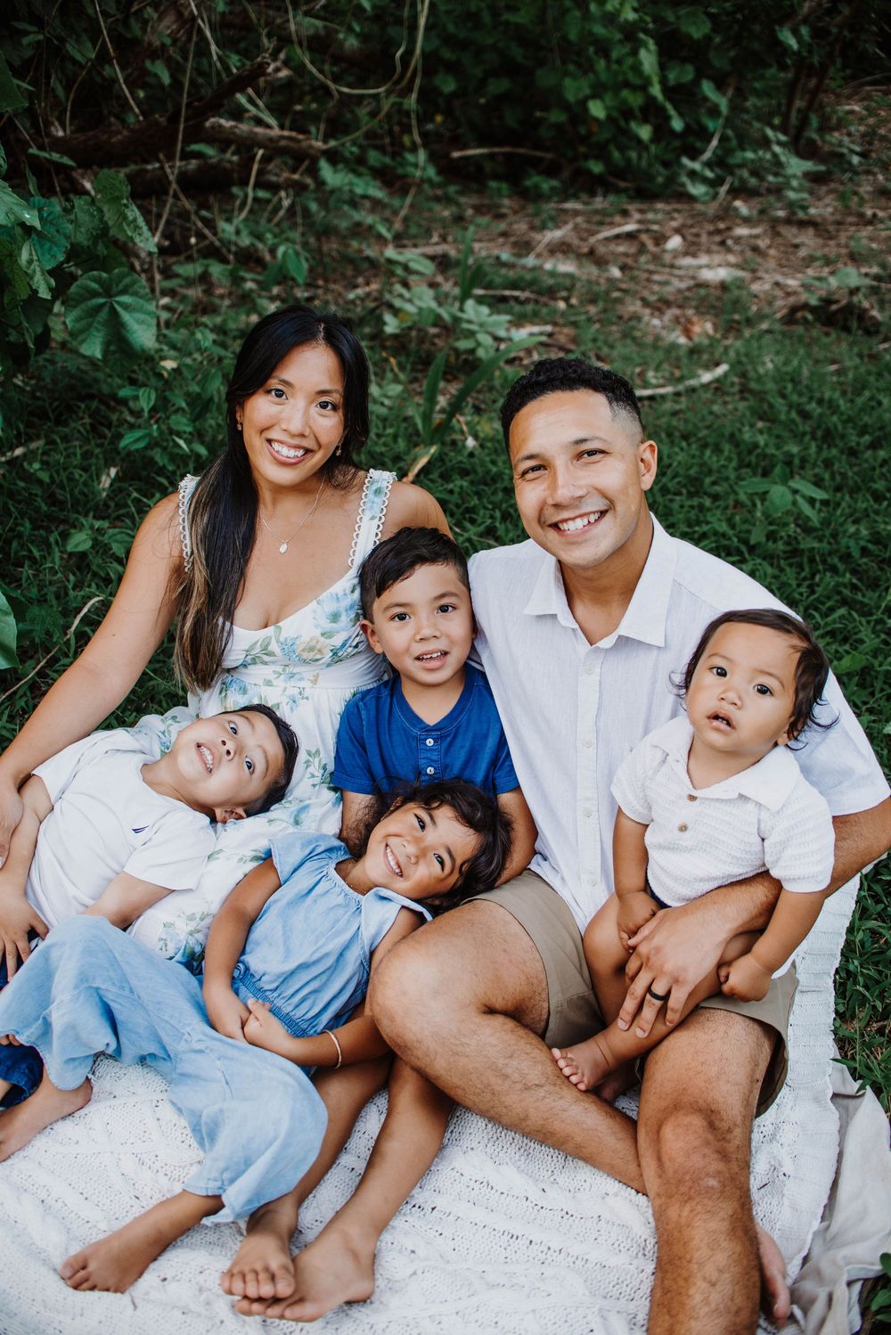 Happy family of five sitting on a blanket outdoors, smiling parents with three young children in a lush green setting.