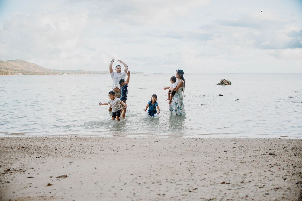 Family playing in shallow ocean waves on a sandy beach with mountains in the background on a cloudy day.