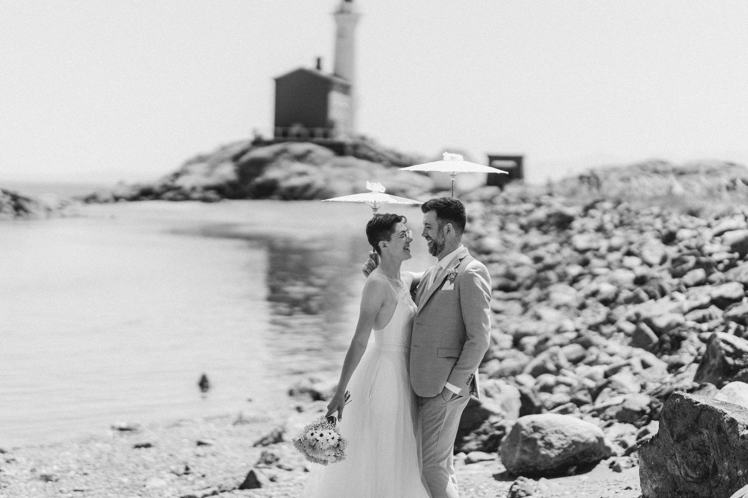 A romantic black and white wedding photo with a lighthouse in the background on a rocky shoreline.