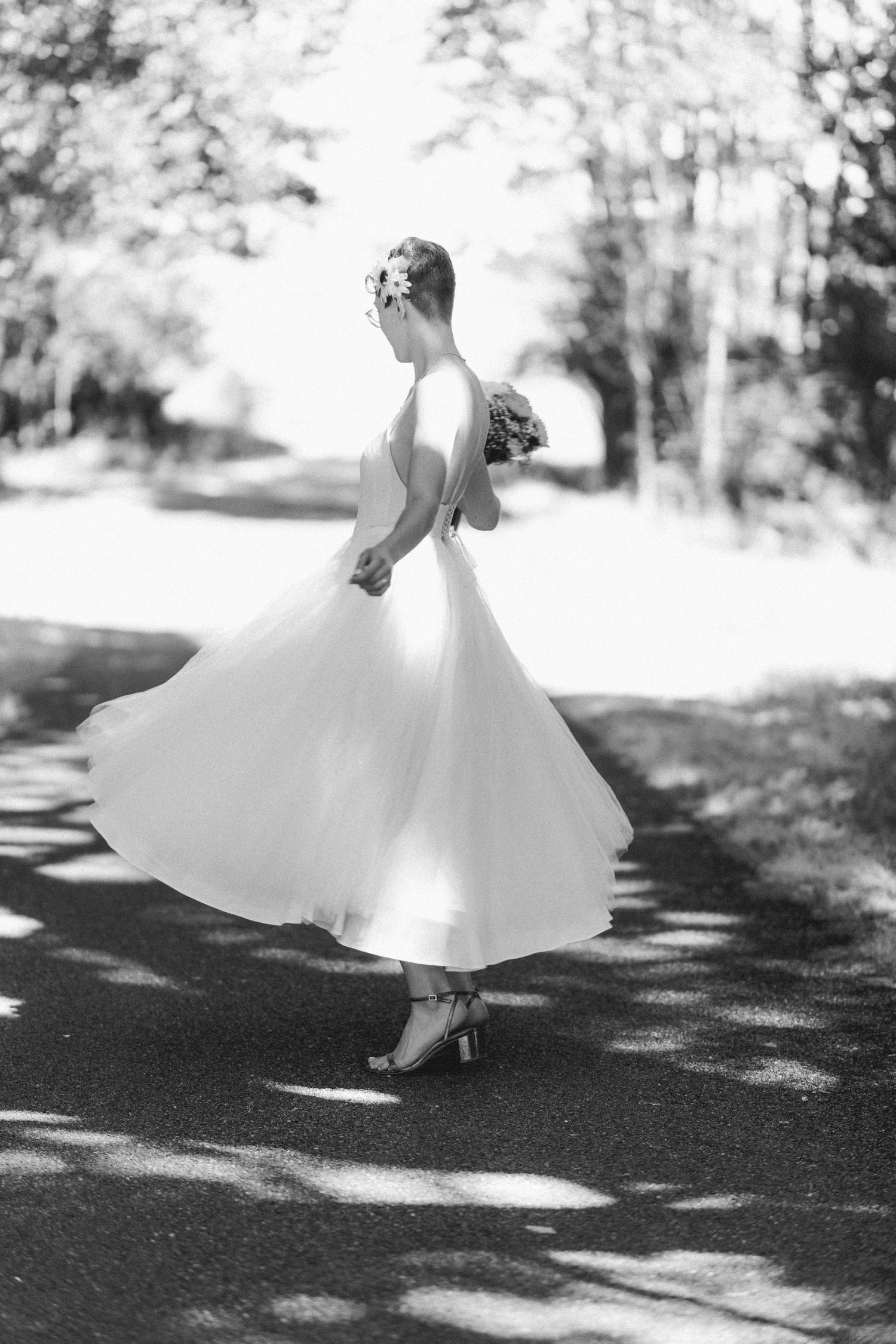 Black and white photo of twirling bride in tea-length dress on tree-lined path.