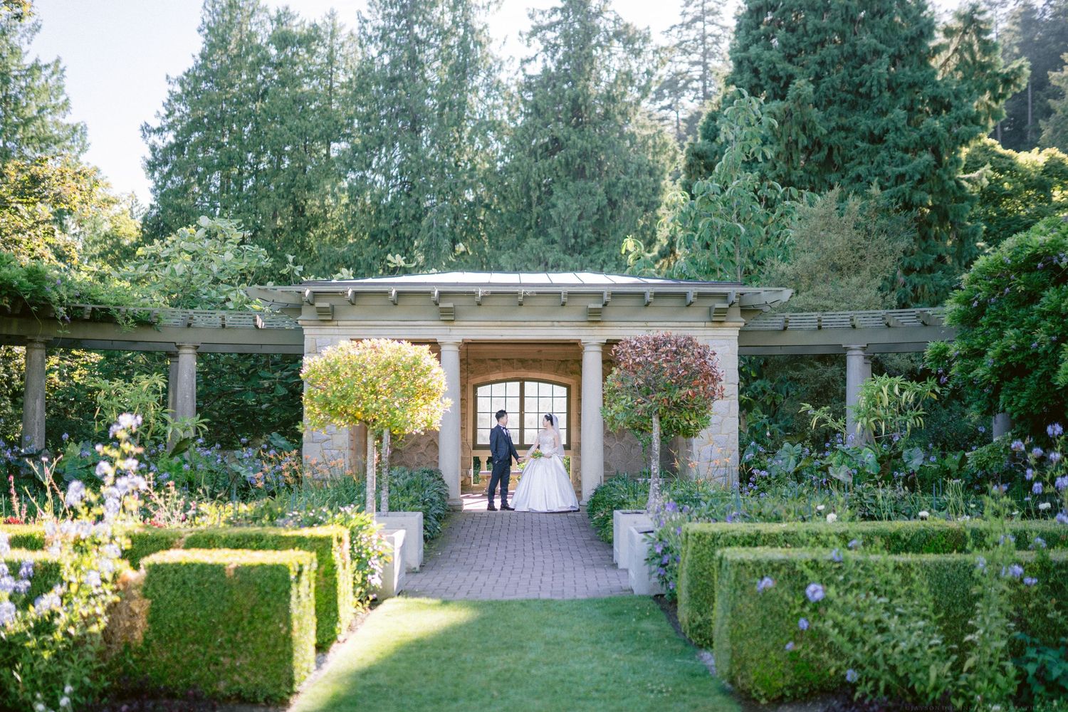 Formal garden path lined with hedges leads to covered archway with hanging planters.