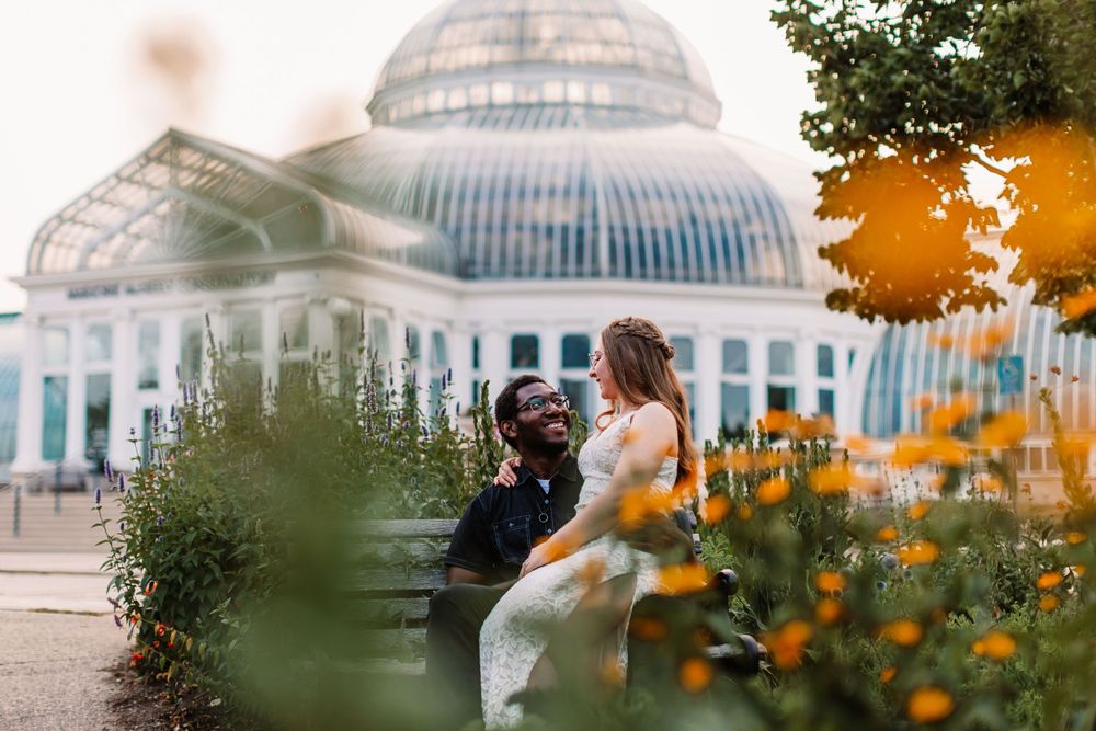 Romantic couple embraces near a conservatory with vibrant orange flowers in the foreground.