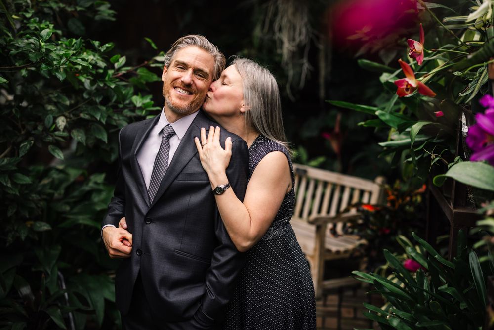 A couple shares an intimate moment in formal attire surrounded by garden flowers.