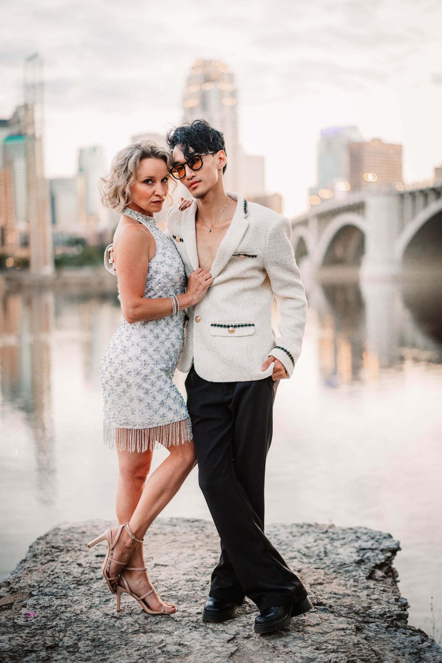 A romantic photoshoot on rocky riverbank with city bridge and skyline in soft focus background.