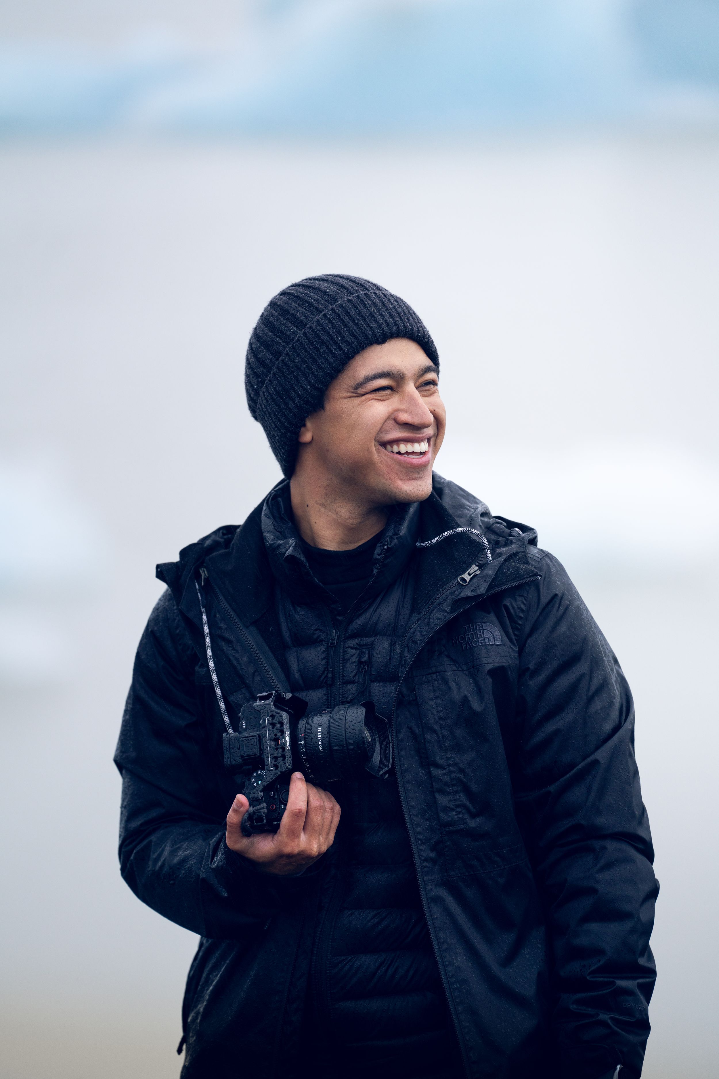 Person in winter gear smiling while wearing black beanie and jacket outdoors.
