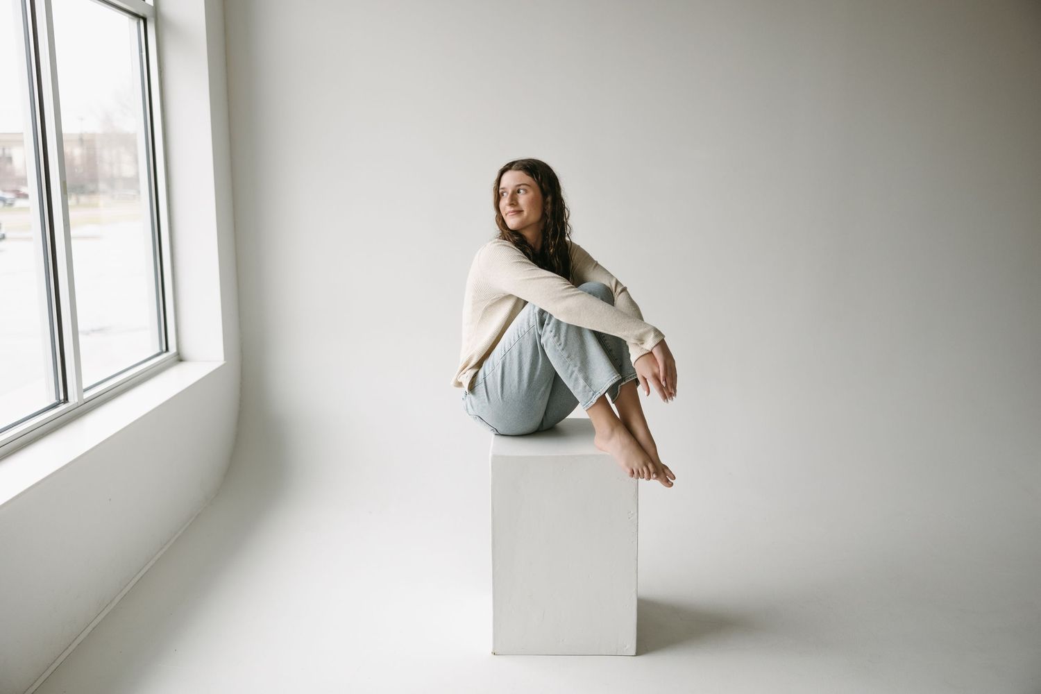 Minimalist portrait of someone sitting on a white cube against a bright window.