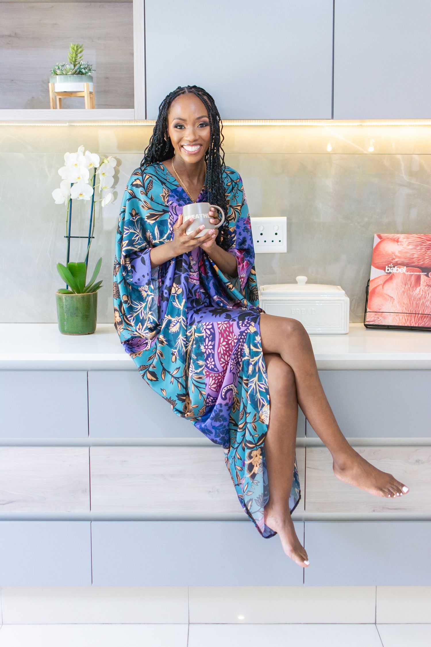 A person in a vibrant blue and purple floral robe relaxes on white kitchen counters with an orchid plant nearby.