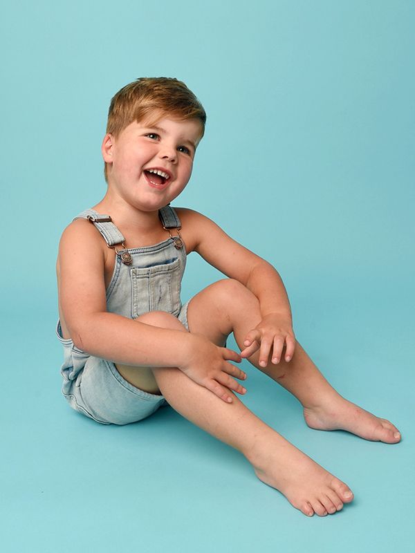A joyful young boy in dungarees sits on a blue background, captured during a summer family photoshoot at Abigail's Studio