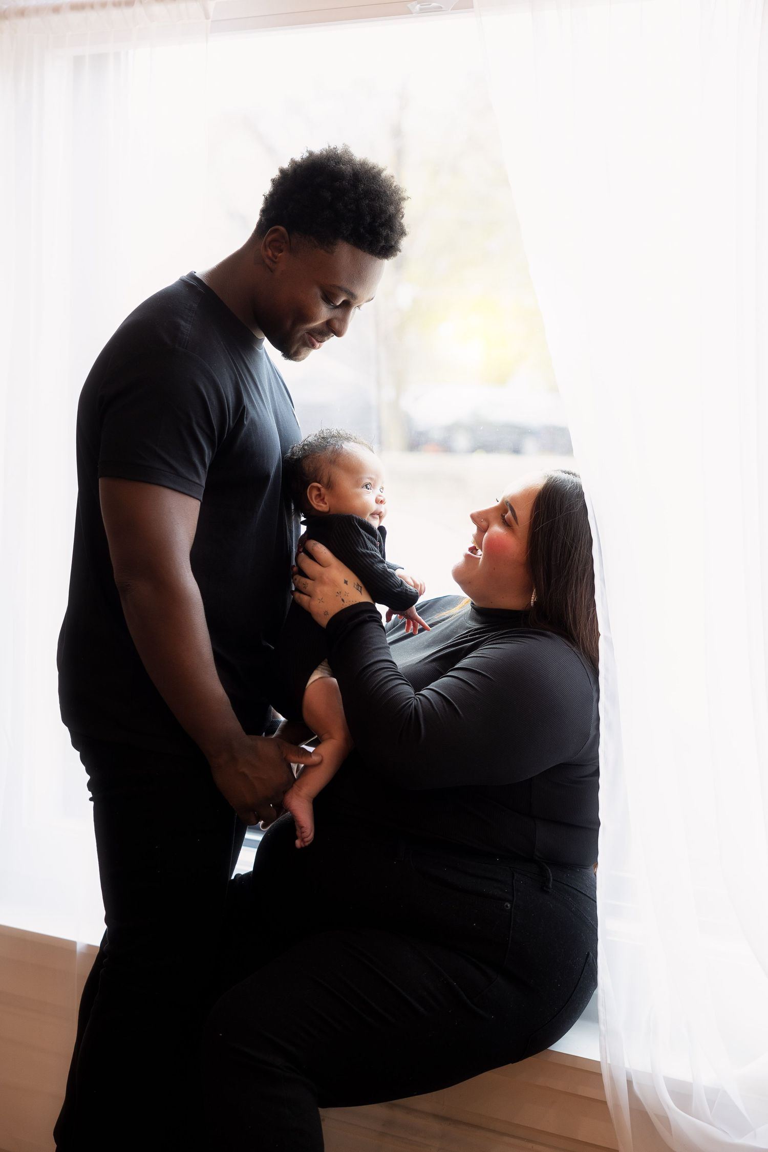 Family shares tender moment near window with baby in black outfits.