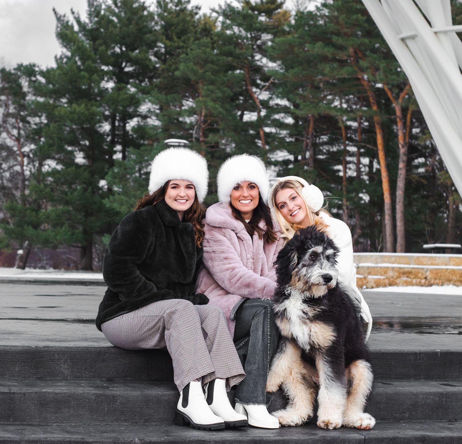 Three women in winter coats pose with Australian Shepherd dog in snowy outdoor setting.