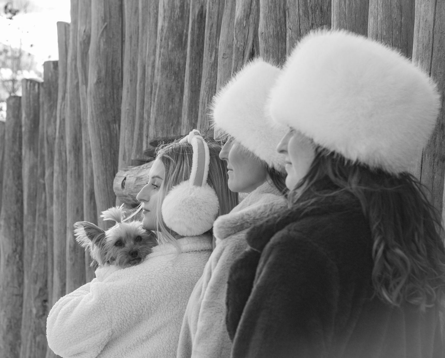 Two people wearing white fur hats looking off into distance against wooden fence in winter scene.