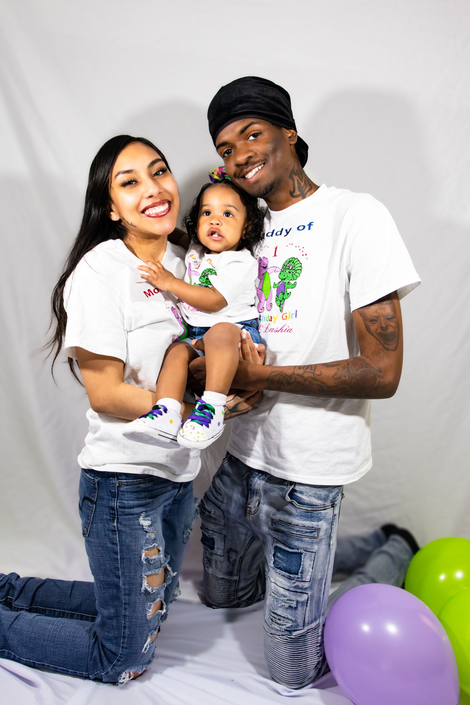 Family in matching white t-shirts poses together with colorful balloons in studio setting.