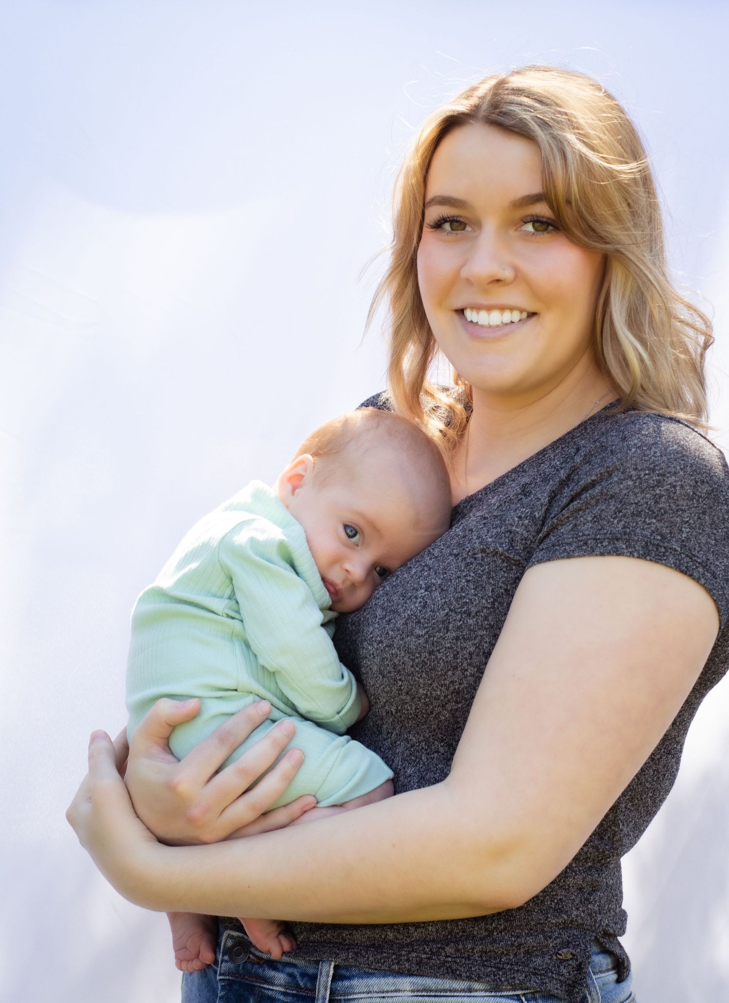 Parent in gray shirt holding infant wearing green outfit against light background.