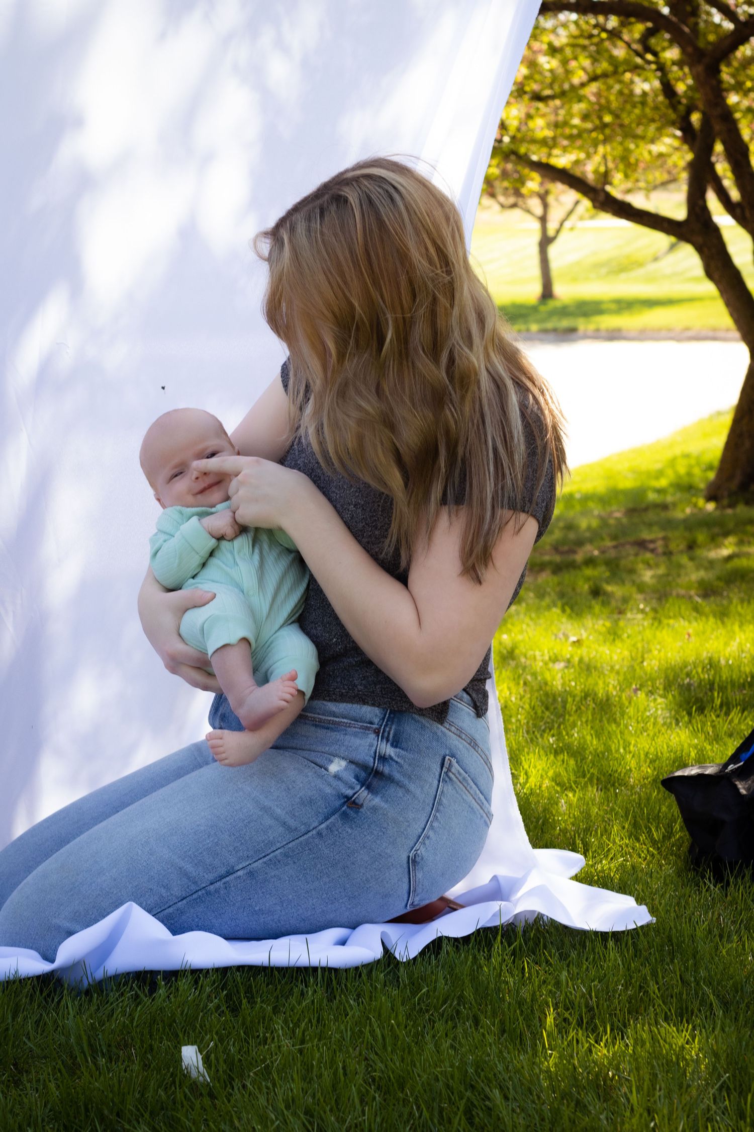 Parent sitting on grass holding baby in mint green outfit on sunny day.