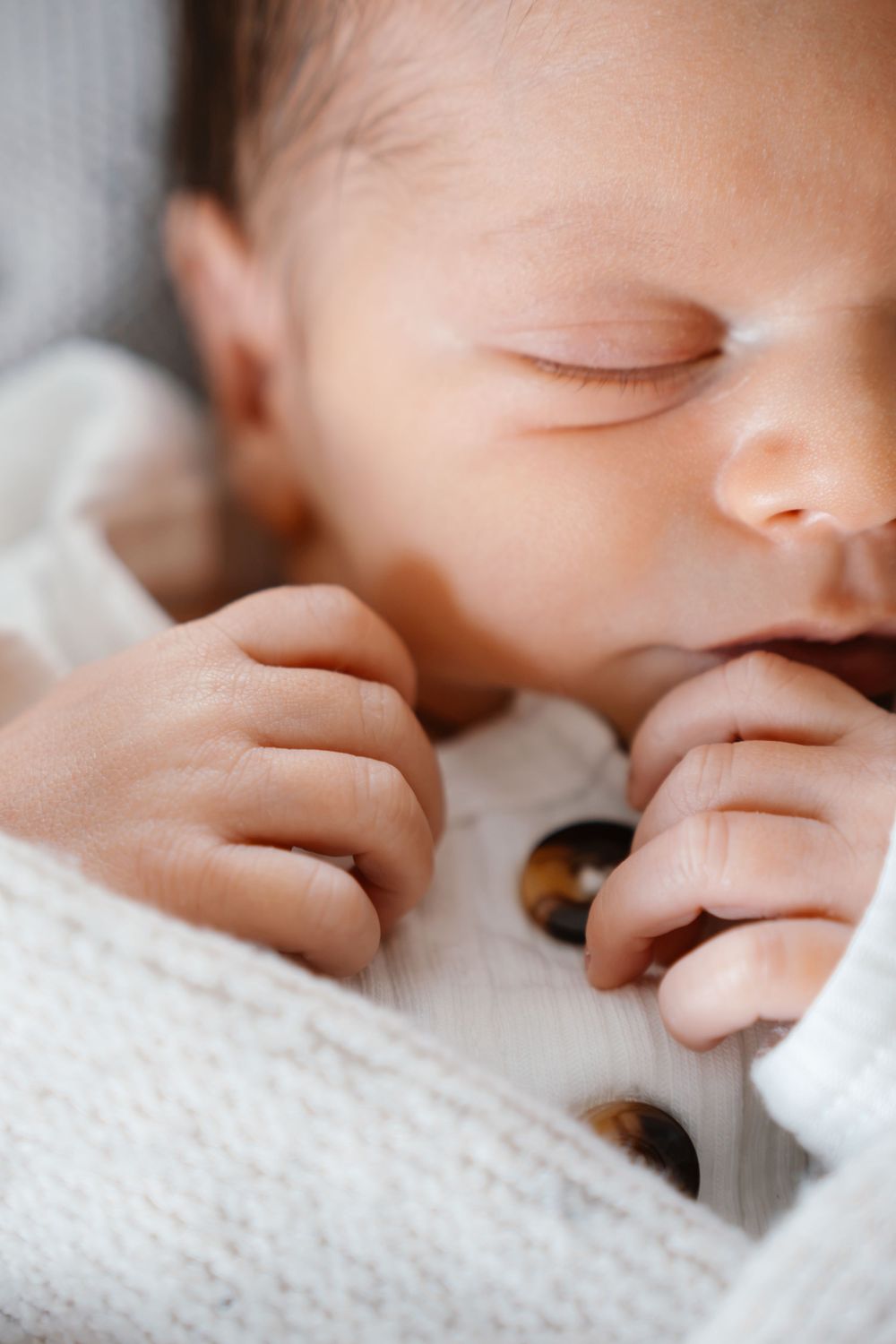 Peaceful newborn baby sleeping in white knit outfit with tiny hands curled near face, close-up portrait photo.
