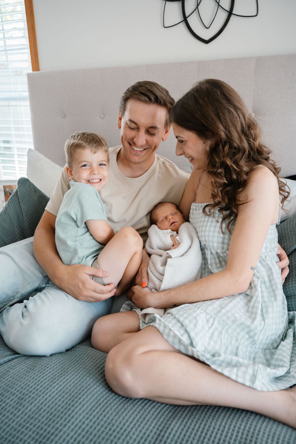 Happy family sitting on bed with toddler son and newborn baby, smiling parents enjoying precious moment together.
