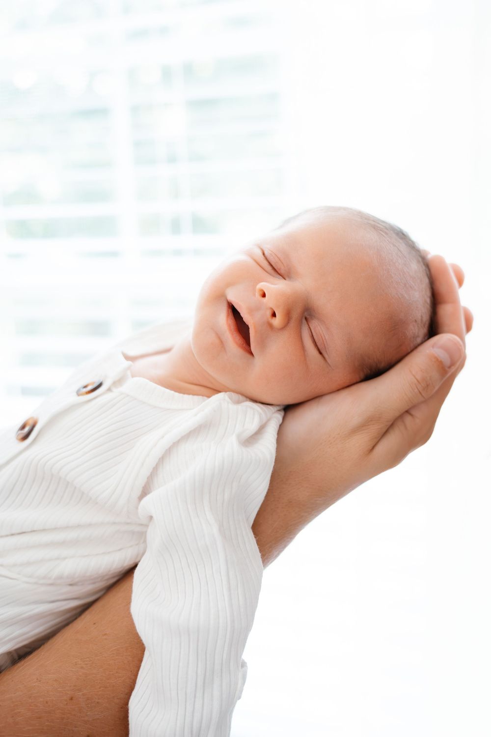 Newborn baby in white onesie cradled in adult hands, eyes closed, smiling peacefully against bright window background.