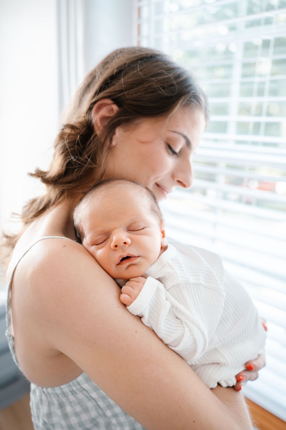 A mother tenderly holds her sleeping newborn baby wrapped in white near a bright window.
