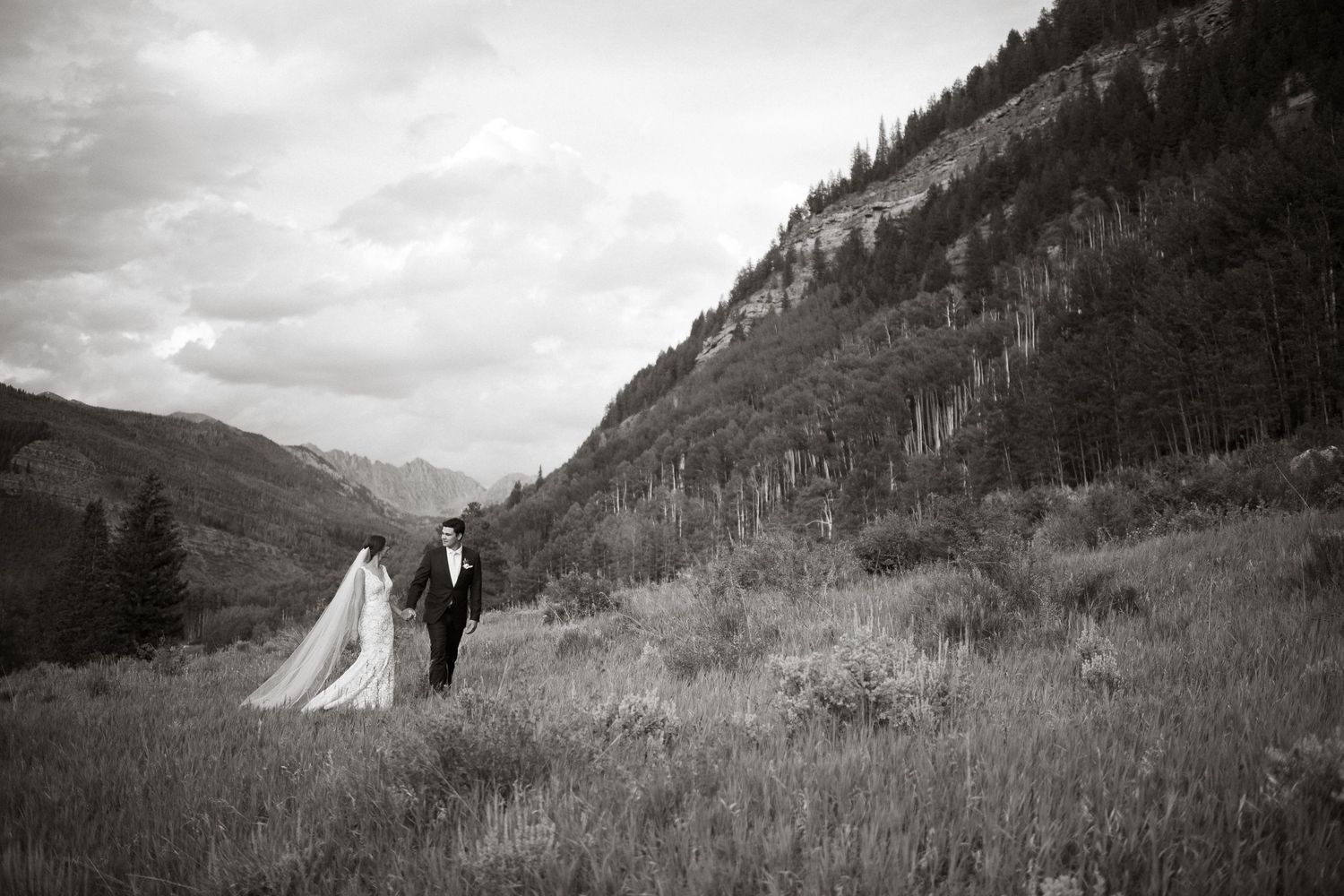 Black and white wedding photo of couple walking through mountain meadow landscape.