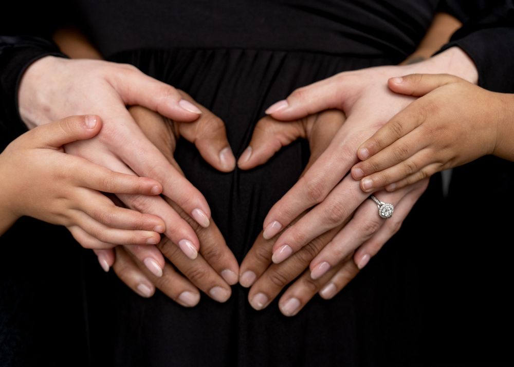 Hands forming a heart shape over a pregnant belly against a black background.