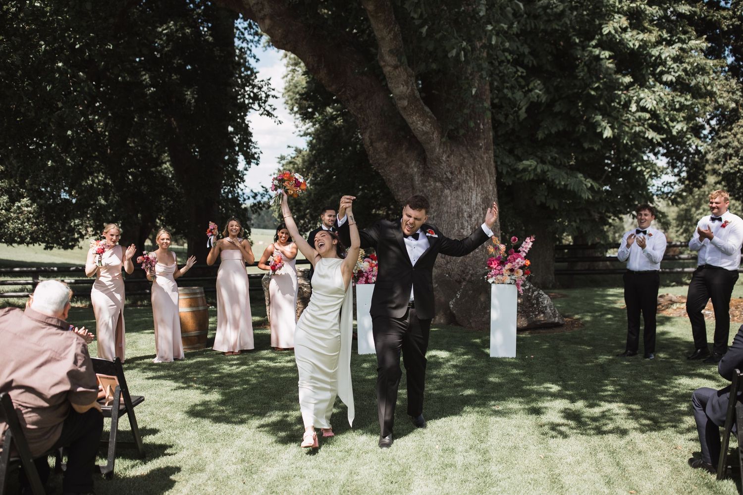 Wedding celebration in a shaded garden setting with guests gathered under trees.