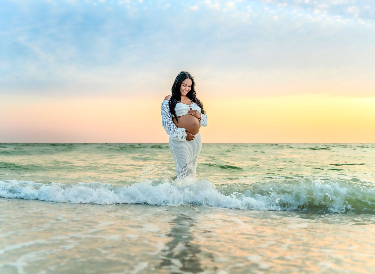 A figure in white stands in ocean waves during a beautiful beach sunset.