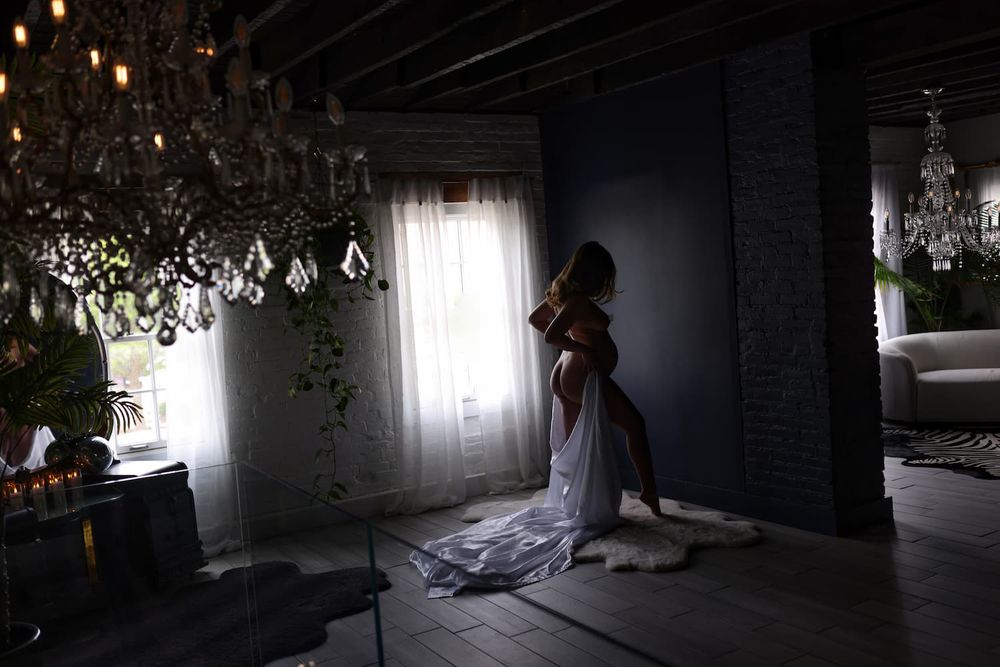 Woman in elegant white gown stands by window in luxurious room with crystal chandelier and moody lighting.