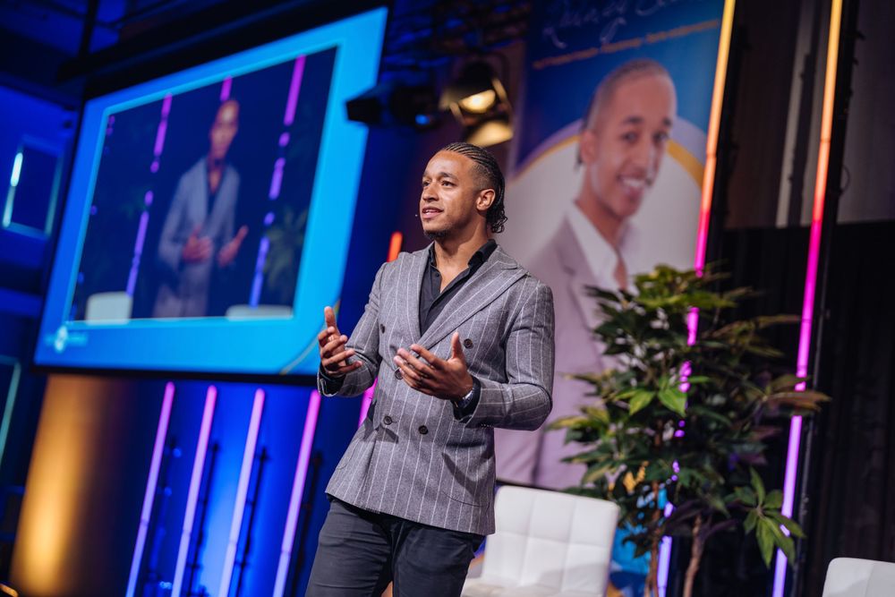 Professional speaker presenting on stage at a conference with blue lighting and digital displays.