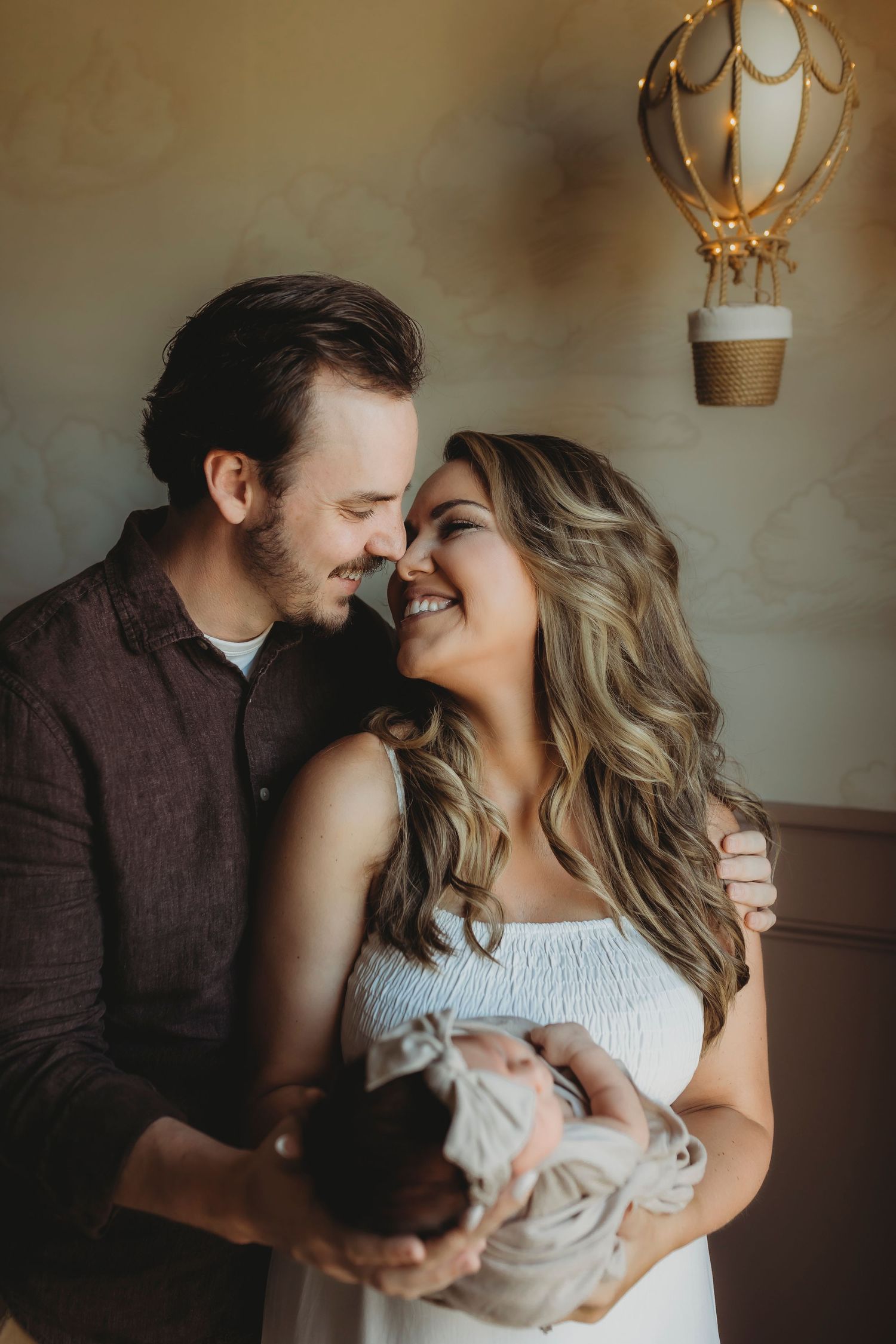 A couple shares an intimate moment while holding their newborn baby under a decorative hot air balloon light.