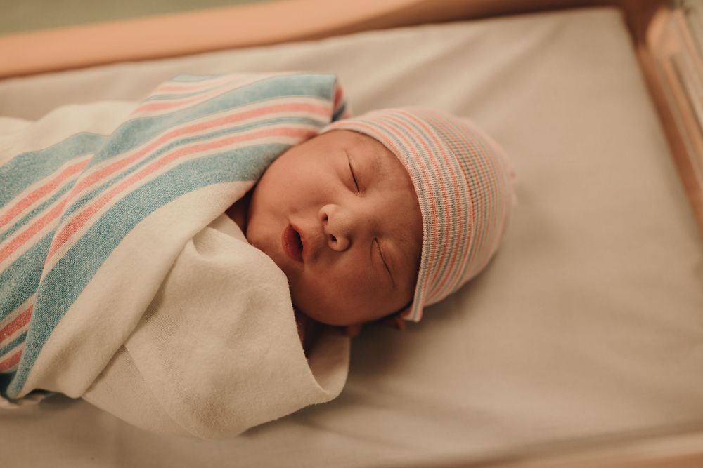 Newborn baby in white and pink striped hospital blanket sleeps peacefully on bed.